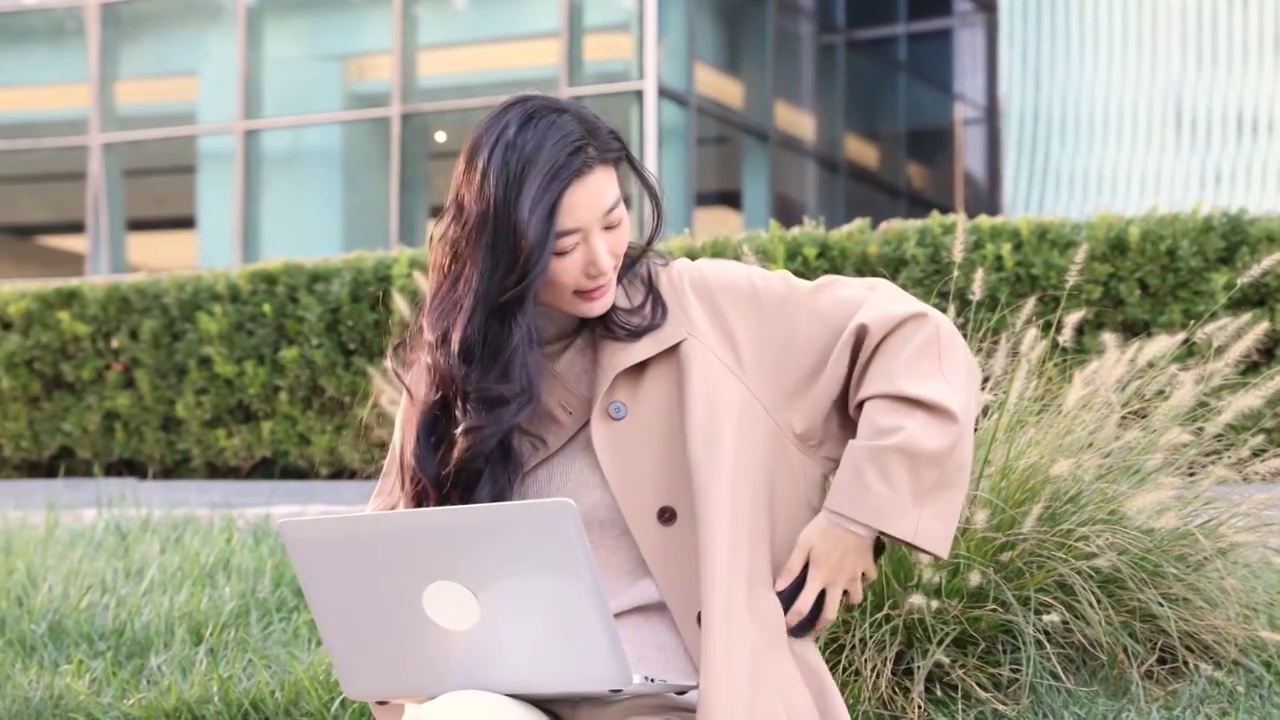 高知女商人在户外办公园区把手机放进风衣口袋后使用笔记本电脑工作视频下载
