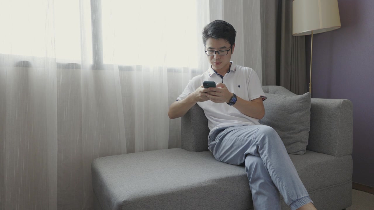 4K年轻男性在窗户旁边沙发上玩手机视频下载