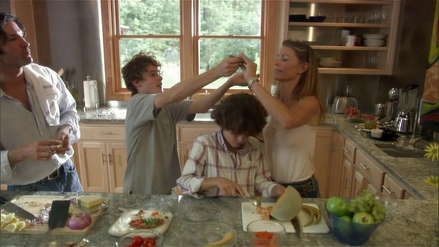 中等镜头的女人帮助男孩从哈密瓜中挖出种子/另一个男孩在厨房给女人吃煎蛋卷/父亲在吃葡萄视频下载