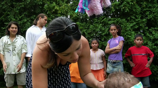 一名女子蒙住小男孩的眼睛，拿着棍子站在皮纳塔下面/背景中的人们/新泽西视频素材