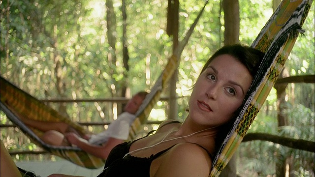中拍女子躺在吊床上听耳机/男子躺在吊床上(背景)/巴西亚马逊视频素材