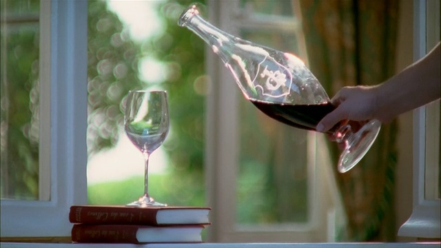 拿起一瓶红酒，把酒倒进一堆书上面的杯子里，把杯子放在杯子旁边视频素材