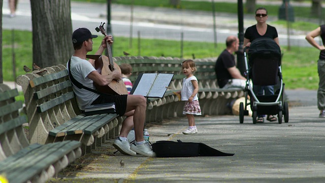 一个男人在中央公园的长椅上弹吉他。两个小孩过来看。视频下载