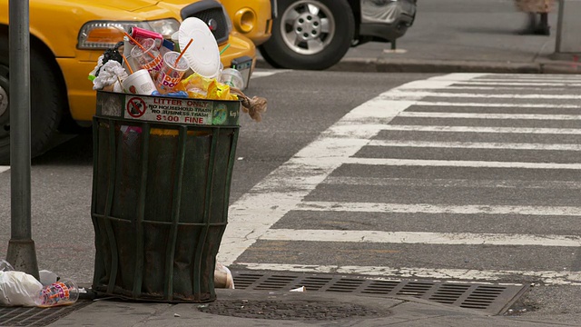 当汽车和行人经过的时候，一个满溢的垃圾桶放在街角。视频下载
