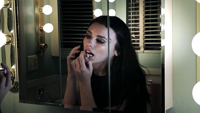 一名年轻女子站在浴室的镜子前，一边讲手机一边涂口红/美国加州棕榈谷视频下载