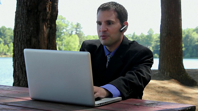 美国波特兰市湖畔附近，MS Man坐在桌子上，一边用蓝牙聊天一边在笔记本电脑上打字视频素材