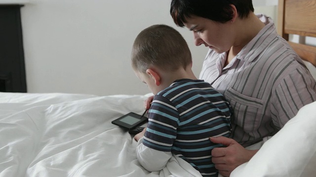 小男孩在床上和妈妈玩他的手握电脑游戏视频素材