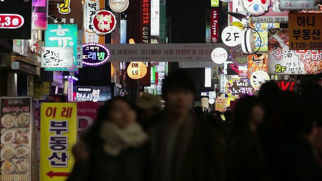 在韩国首尔仁寺洞区，一对LS夫妇在夜晚走过一条布满灯光的酒吧和餐馆标识的街道视频素材