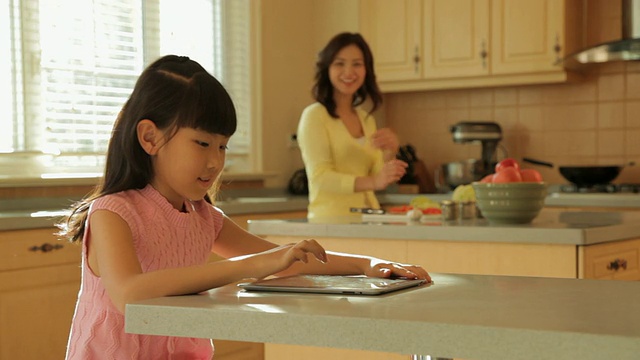 MS SELECTIVE FOCUS女孩在厨房柜台上使用数字平板电脑，母亲在后台做饭/中国视频素材