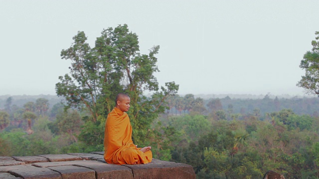 柬埔寨吴哥窟(暹粒)的一座古庙上，一名佛教僧侣正在冥想，另一名僧人站在丛林中望着太阳升起视频下载