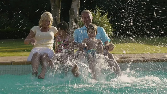 孙辈和祖父母坐在泳池边把脚溅到水里的慢动作。视频素材