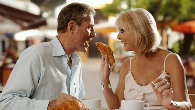 老夫妇坐在咖啡馆吃牛角面包，一起大笑。视频下载