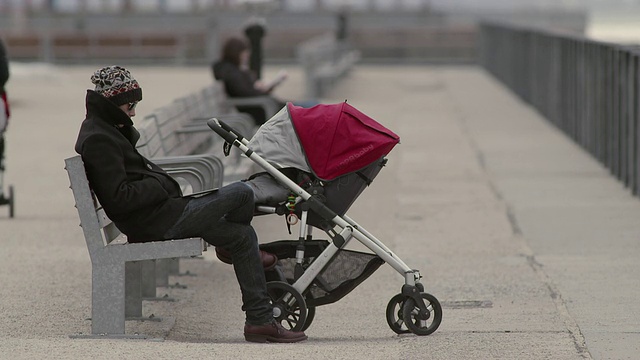 一个男人静静地坐着，旁边的马车里有一个睡着的婴儿。一个灰色的日子，他们在布鲁克林的一个码头公园。视频素材