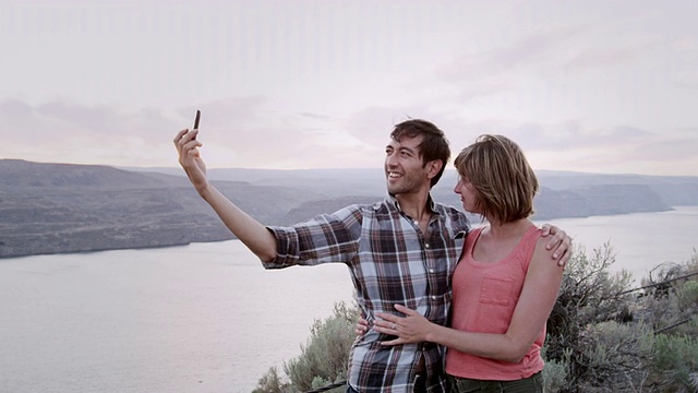 一对情侣站在一条乡村公路边，用智能手机以峡谷为背景拍照视频素材