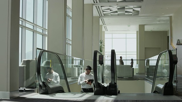 商人使用数码平板电脑，爬上自动扶梯，然后在办公室大厅里穿梭。视频下载