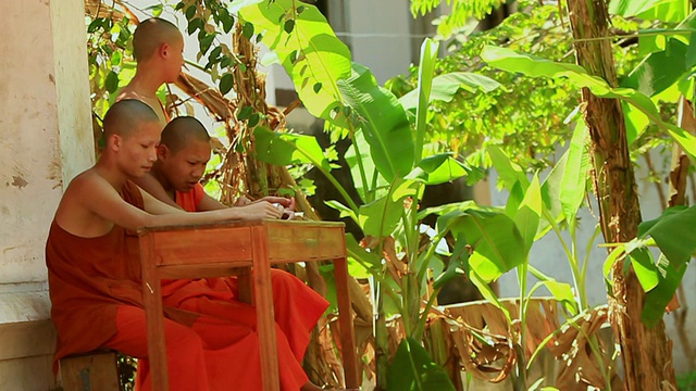 老挝琅勃拉邦，两名身穿藏红花袈裟的年轻和尚坐在长凳上，另一名和尚站着视频下载