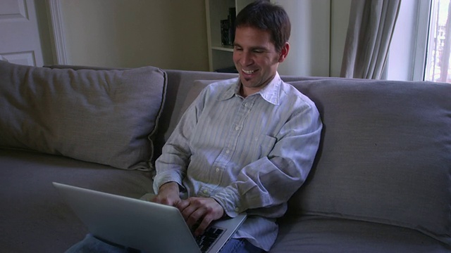 美国纽约，一男子坐在公寓沙发上用笔记本电脑兴奋地打字视频素材