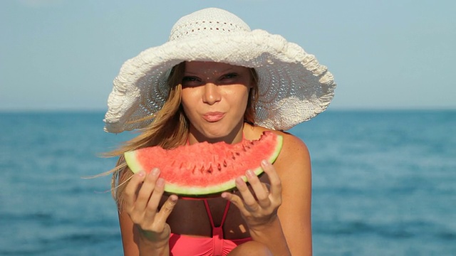 迷人的金发女人戴着帽子在海滩上吃西瓜视频下载