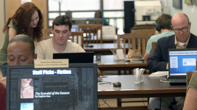 在公共图书馆的阅览室里，一名年轻女子走向一名年轻男子，而其他读者则在那里用耳机阅读或听音乐视频下载