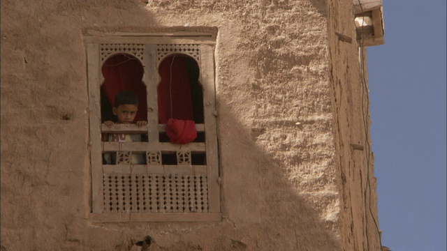 一个小男孩从泥砖公寓楼的装饰窗向外看。视频下载