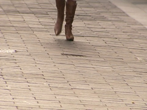 一个女人穿着低垂的靴子和裙子走过人行道在许多行人。视频素材