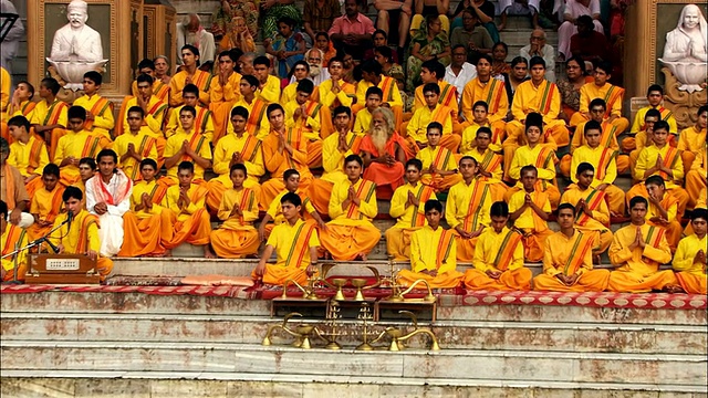 长者坐在一群穿着橙色长袍的男人中间，在恒河边的台阶上祈祷。视频下载