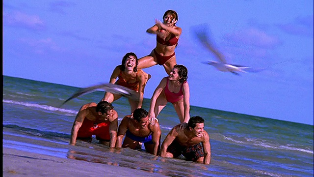 穿着泳衣的人在海滩上形成人体金字塔/他们在拍摄结束时掉下来视频素材