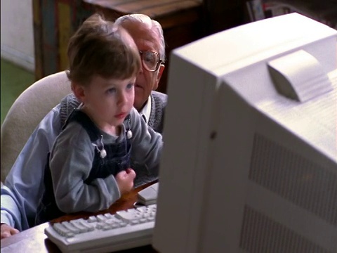 小男孩坐在爷爷身边，一边用电脑+一边指着显示器视频下载