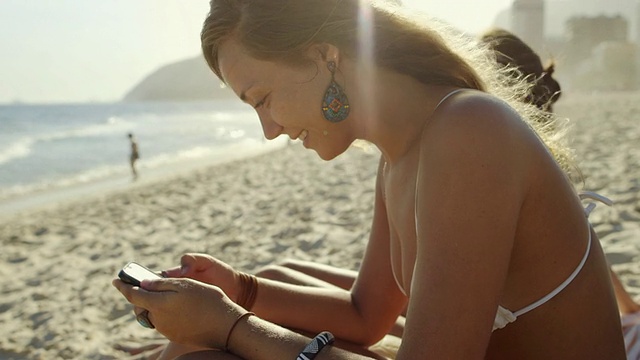 在巴西里约热内卢的伊帕内玛海滩上，一名年轻女子在她的智能手机上打字视频下载