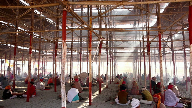 阳光斜射穿过摇摇欲坠的木制脚手架，虔诚的信徒坐在下面的火堆旁，参与宗教仪式。Kumbh梅拉、印度视频下载