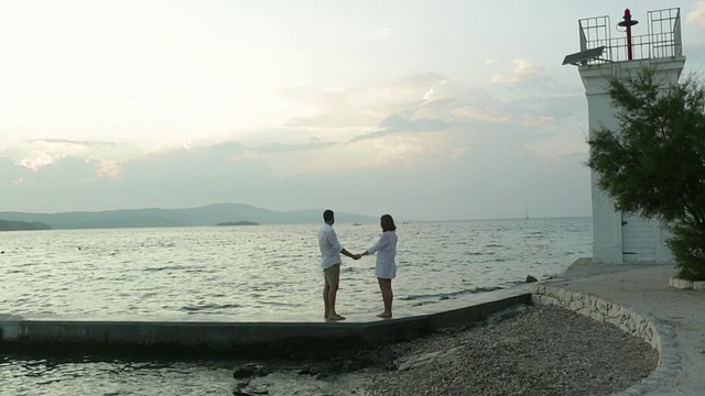 一个年轻人牵着一个女人的手走在海边的一个码头上，他们停下来拥抱并看风景。视频下载