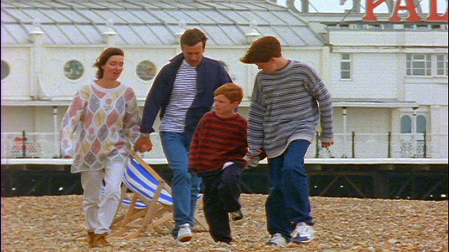 慢镜头一家人在风中漫步在布莱顿海滩上/背景是维多利亚宫码头/英国视频下载