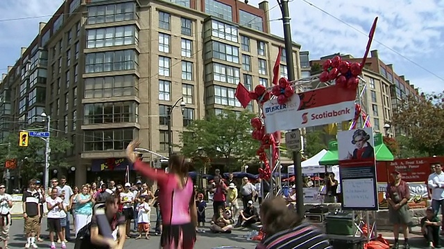 加拿大安大略省多伦多市街头艺术节上，一群人看着两名男子举着弹力梁，而一名女子在空中表演杂技视频素材