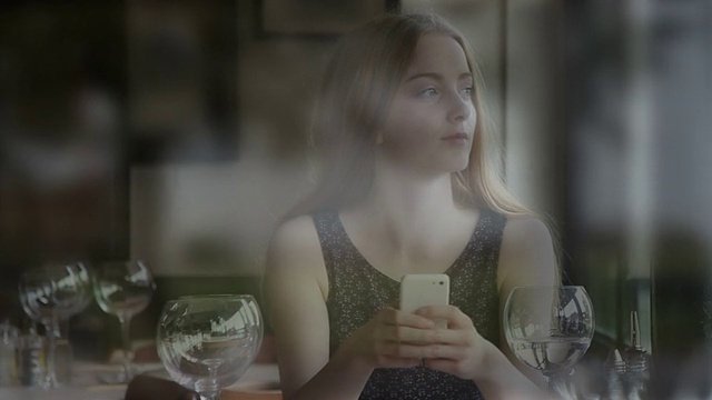 年轻女子在餐厅里用智能手机透过窗户看外面。视频下载
