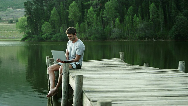 一名男子坐在湖边的码头上使用笔记本电脑视频素材