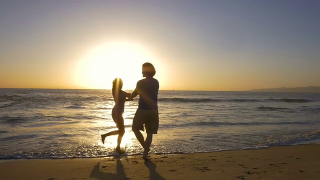 在美丽的海滩上，年轻的夫妇手牵着手，在夕阳下旋转。视频素材