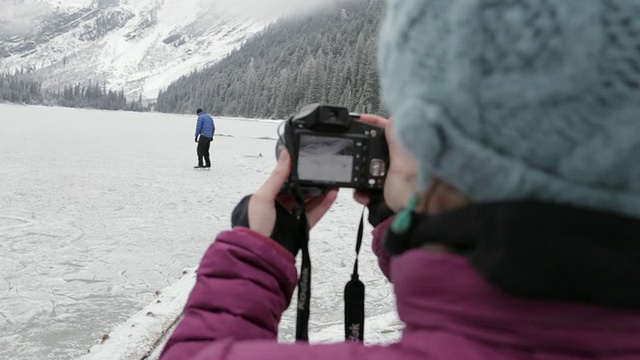 在蒙大拿州冰川公园，一名女子正在拍摄一名男子在结冰的山地湖泊上滑冰的照片视频素材