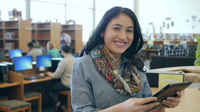 一名西班牙妇女在图书馆里使用数码平板电脑，抬头对着相机微笑视频素材
