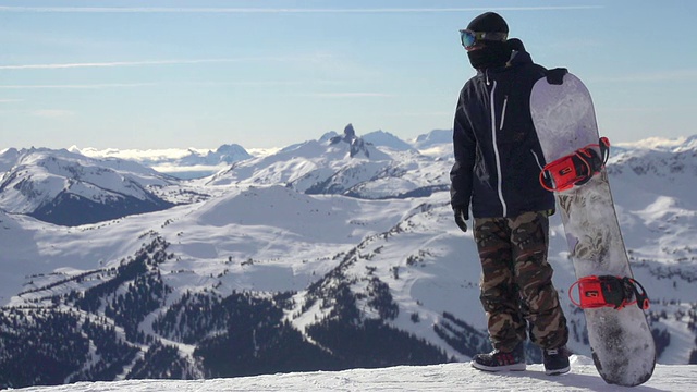 一个年轻人带着滑雪板站在白雪覆盖的山上。——慢动作视频下载