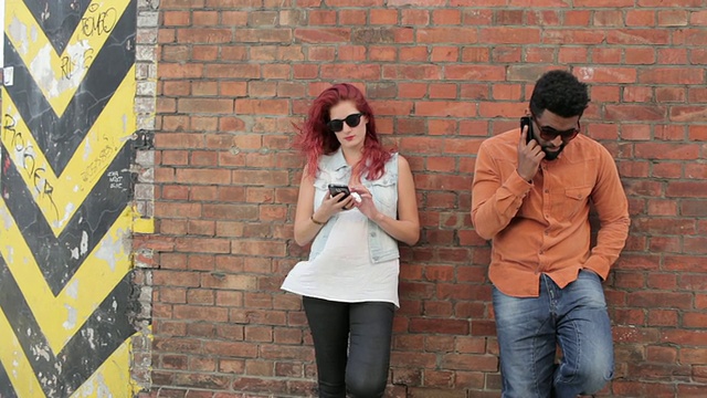 一对情侣靠在外面的砖墙上，看手机，发短信，打电话视频素材