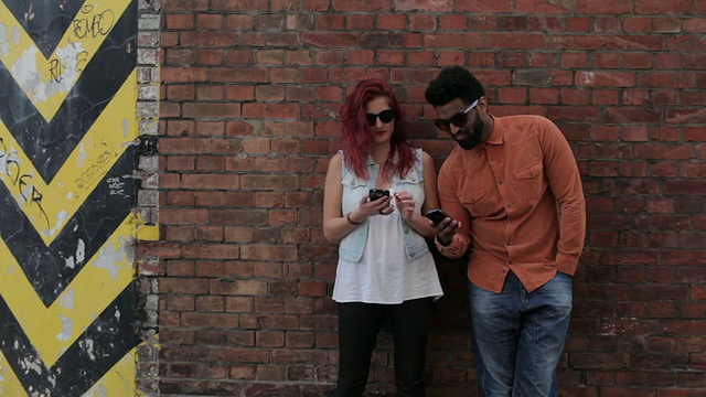 一对情侣靠在外面的砖墙上，看着手机，发短信，互相交谈视频素材