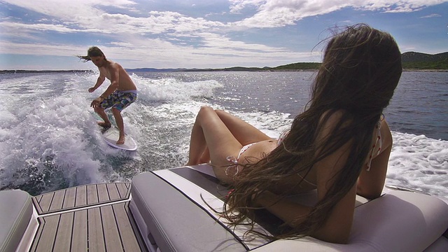 一个年轻人在船后冲浪，一个女人在晒日光浴视频素材