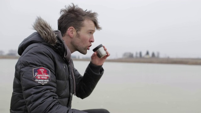 在美国蒙大拿州的乡下，一个年轻人在结冰的池塘上喝着热茶。视频素材