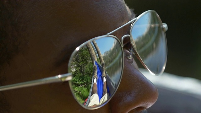 这是一个戴着墨镜的男人躺在山湖附近的吊床上的特写镜头。——慢动作视频下载