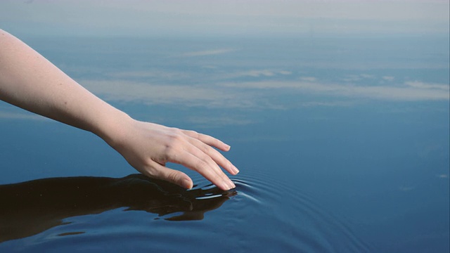 一位妇女用手在蓝天倒映的水中画出涟漪。视频素材