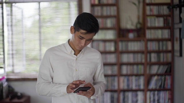 一名年轻男子坐在书架前用智能手机发短信视频素材