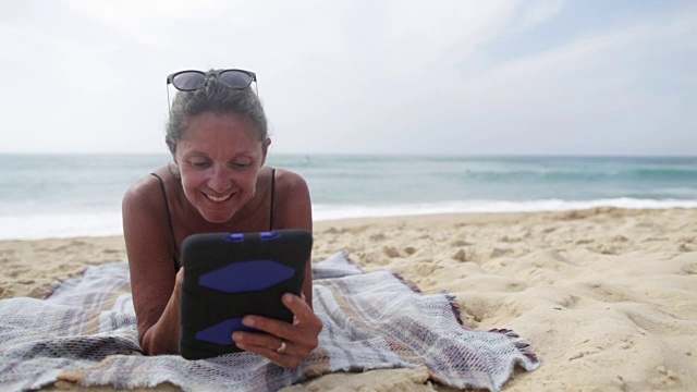 在法国南部的海滩上，一个成熟的女人躺在平板电脑上看书。视频下载