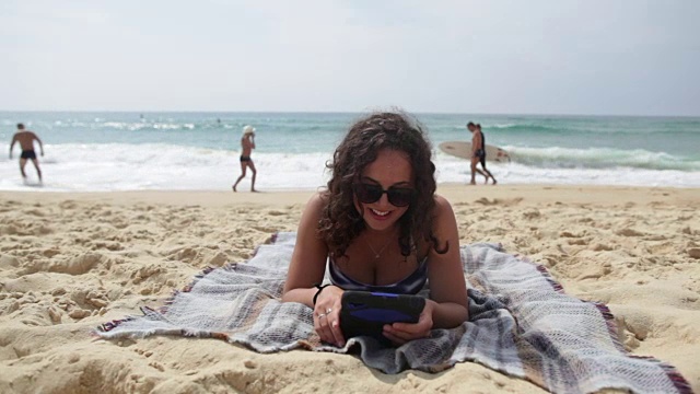 一个年轻女子躺在法国南部的海滩上，在平板电脑上看书，笑着。视频素材