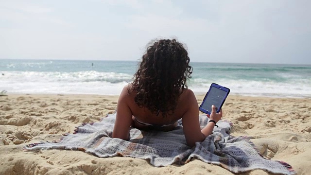 一名年轻女子躺在法国南部的海滩上阅读平板电脑。视频下载