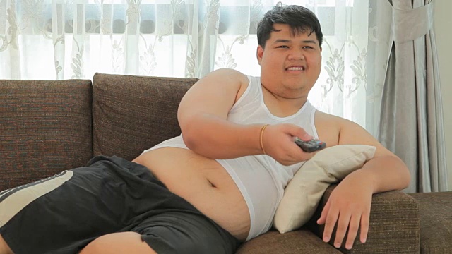 一个超重的亚洲人坐在沙发上，手里拿着遥控器想看电视视频素材
